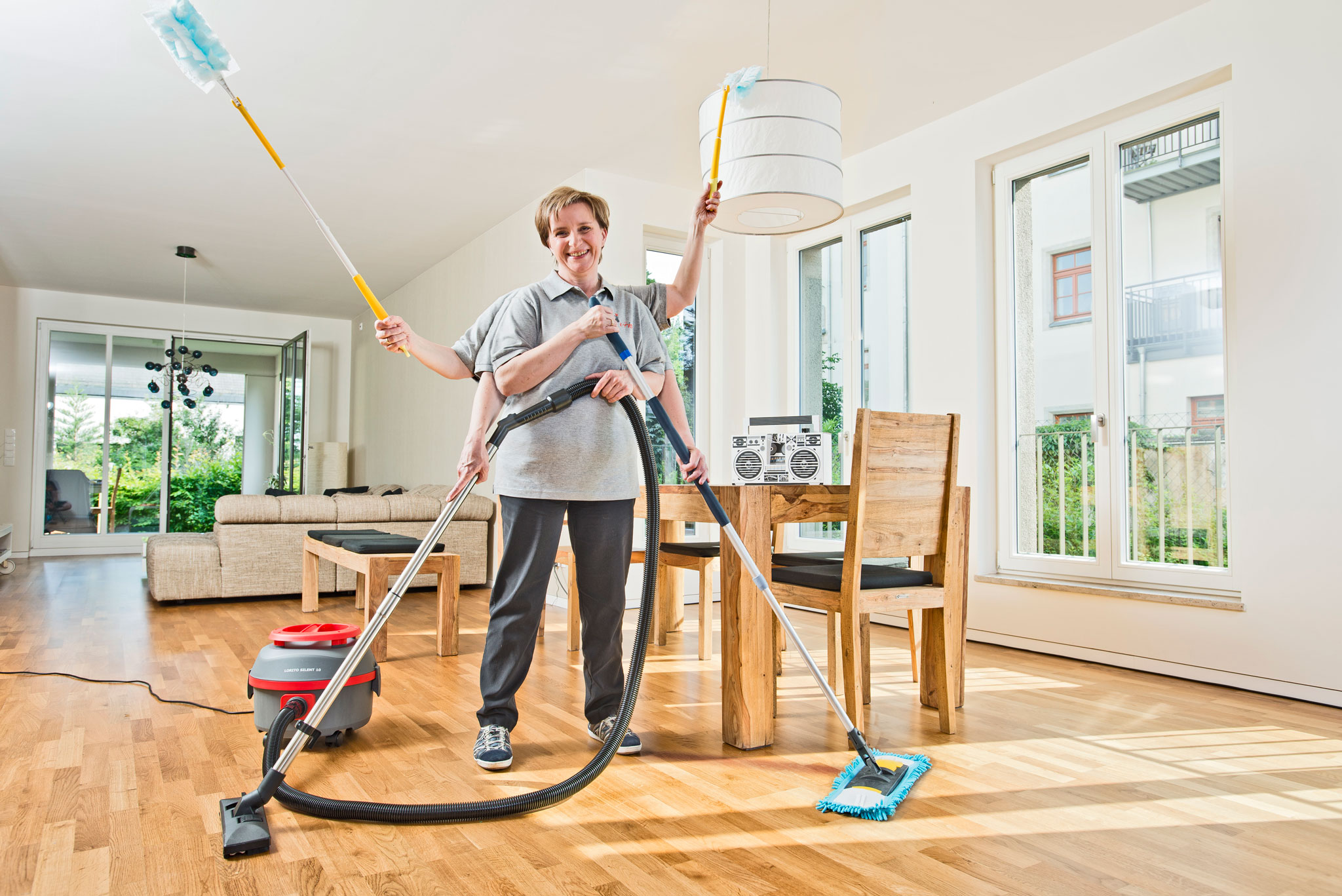 Eine lächelnde puris Haushaltsreinigerin in Dresden beweist mit ihren sechs Armen wahre Multitasking-Stärke: Sie staubwischt gleichzeitig die Decke und die Lampe, saugt den Boden mit einem Staubsauger und wischt mit einem Mopp – alles auf einmal, in einem hellen, modernen und sauberen Wohnzimmer, mit einem Radio auf dem großen Esstisch im Hintergrund. Sie lächelt strahlend und hat sichtlich Freude an ihrer Arbeit.
