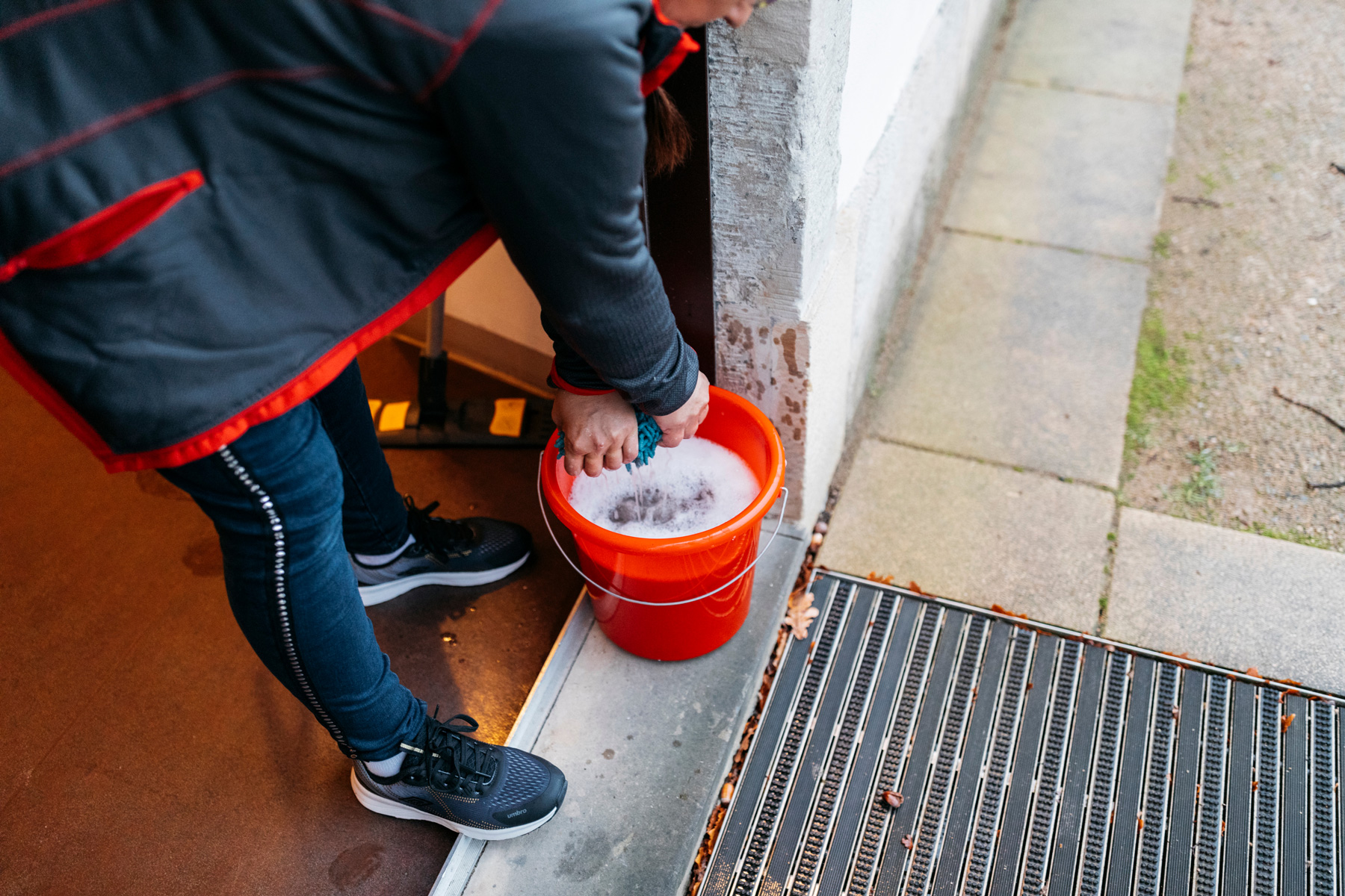 Eine zuverlässige Reinigungskraft der puris Haushaltsreinigung in Dresden trägt Arbeitskleidung mit roten Akzenten und kniet an einem Hauseingang, um einen Lappen in einem roten Eimer mit Seifenwasser auszuwringen.